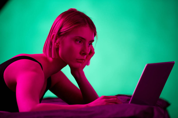 Blondes ernstes Mädchen in schwarzer Unterwäsche schaut auf Laptop-Display, während Netzwerk oder Online-Video auf dem Bett am Morgen oder Abend - Foto, Bild