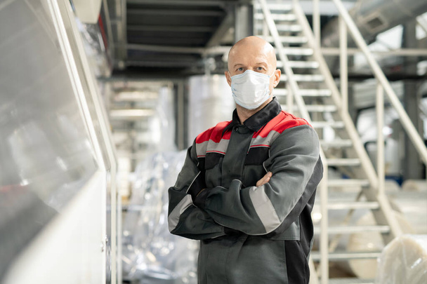 Bald mature worker of contemporary large polymer processing factory in workwear and protective mask crossing arms by chest in warehouse - Foto, afbeelding