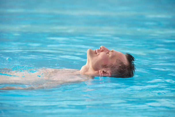 Kopf des glücklichen jungen entspannten Mannes hält die Augen geschlossen und genießt das warme Wasser des Schwimmbades im modernen Kurzentrum - Foto, Bild