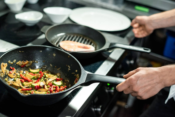 Руки шеф-повара мужского пола жарят кусок лосося и овощное рагу на сковородках, стоя у электрической плиты и готовясь здоровый обед - Фото, изображение