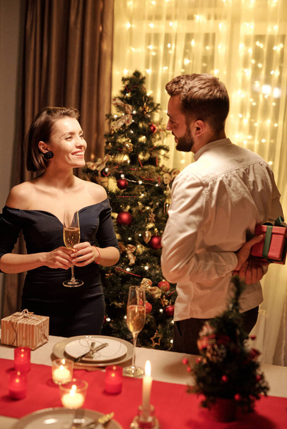 Joven morena elegante hembra con flauta de champán mirando a su marido escondiendo caja de regalo con Navidad presente detrás de la mesa festiva - Foto, Imagen