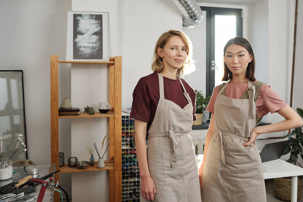 Dwie odnoszące sukcesy młode międzykulturowe rzemieślniczki w ubraniach roboczych stojące blisko siebie przed kamerą w dużym warsztacie lub pracowni - Zdjęcie, obraz