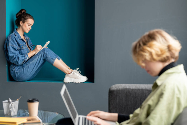 Contemporânea jovem morena empresária em jeans casualwear sentado contra a parede e escrevendo novas ideias para o projeto no notebook - Foto, Imagem