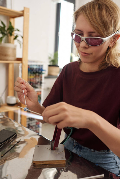 Young female artisan in protective eyeglasses and casualwear decorating glass workpiece on stick with melted paint while holding it over table - 写真・画像