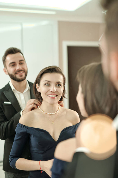 Happy young beautiful woman in black evening gown standing in front of mirror and looking at her elegant husband helping with necklace - Fotó, kép