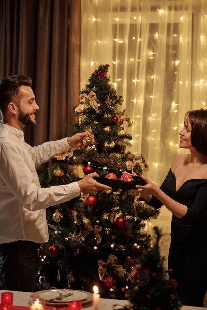 Happy young brunette woman in elegant black dress giving toy balls to her cheerful husband putting decorations on Christmas tree at home - Photo, Image