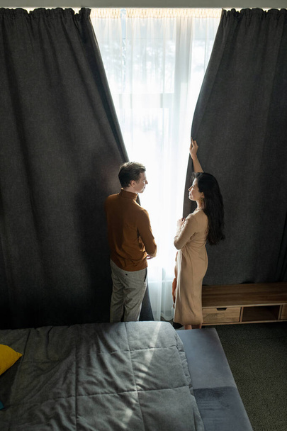 Nuori mies ja hänen vaimonsa seisovat sängyn ja ikkunan välissä katsellen toisiaan ja vetävät pimeitä verhoja hotellihuoneeseen illalla. - Valokuva, kuva