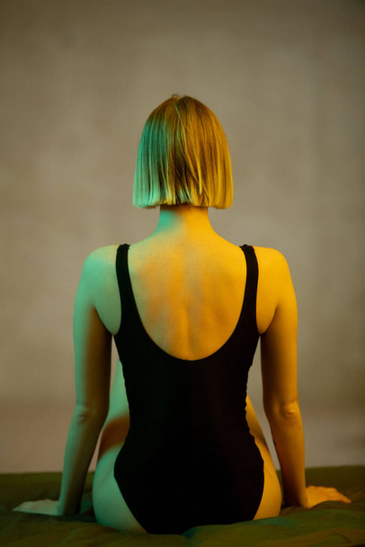 Rear view of young blond slim woman in black leotard sitting on bed in front of camera against grey wall in bedroom or studio - Photo, Image