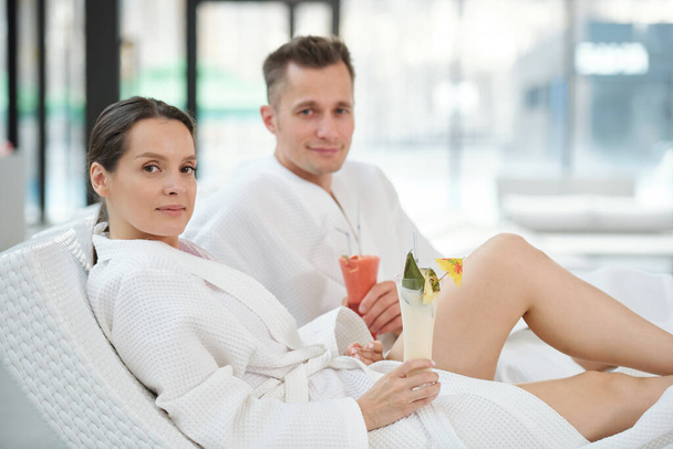 Jeune couple en peignoirs blancs se détendre dans le centre spa moderne et avoir des cocktails tropicaux tout en étant assis sur des chaises longues devant la caméra - Photo, image