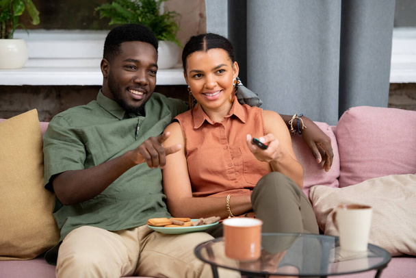 Happy teenage boy embracing his mixed-race girlfriend with remote control while both sitting on couch in front of tv set in living-room - Photo, Image