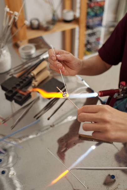 Hands of young female professional lampworker burning glass workpiece with fire while holding it over burner in workshop or studio - Zdjęcie, obraz