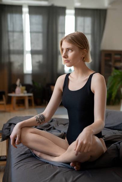 Blond girl in black leotard crossing legs while sitting on bed against window and bedroom interior and thinking of plans for new day - Photo, Image