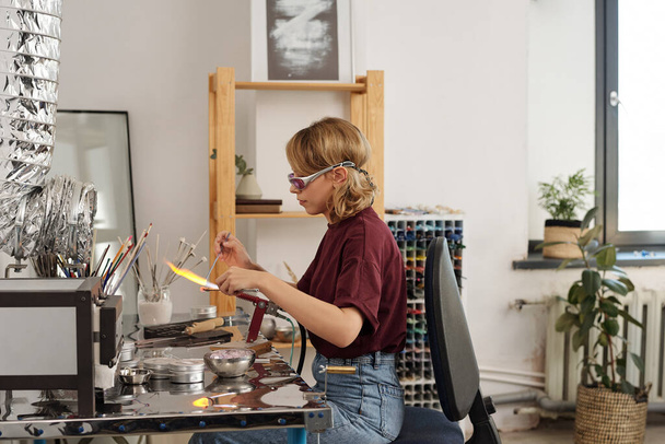 Молодая женщина в повседневной одежде и защитные очки с помощью горелки, сидя за столом в мастерской и держа заготовку над огнем - Фото, изображение