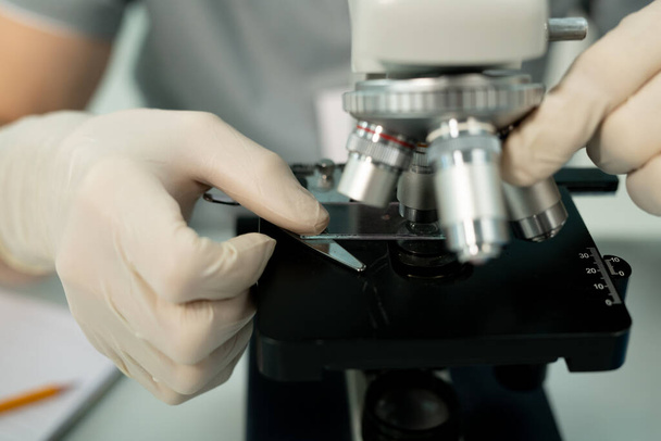 Mão enluvada de funcionário contemporâneo de laboratório científico que põe o vidro com a amostra química pela lente do microscópio durante a investigação - Foto, Imagem