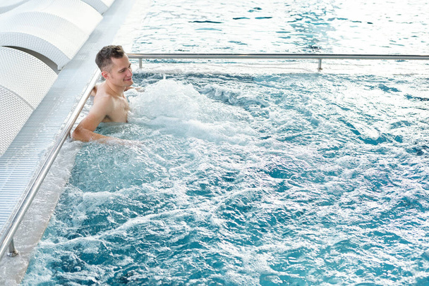 Joven hombre activo disfrutando de la sensación de burbujeante agua mientras está de pie junto al borde de la piscina con una fila de tumbonas blancas detrás - Foto, Imagen