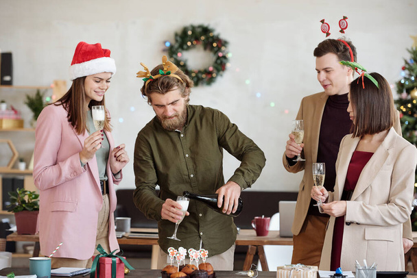企業のパーティー中にクリスマスのお祝いの間に同僚の笛にシャンパンを注ぐ幸せな若いひげを生やしたビジネスマン - 写真・画像