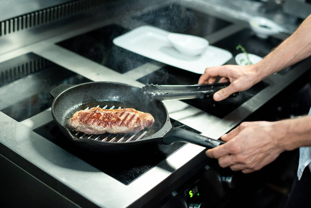 Mains de chef avec pinces de cuisine métalliques sur le bord de la poêle à frire chaude avec morceau de viande rôtie debout près de la cuisinière électrique et la cuisson - Photo, image