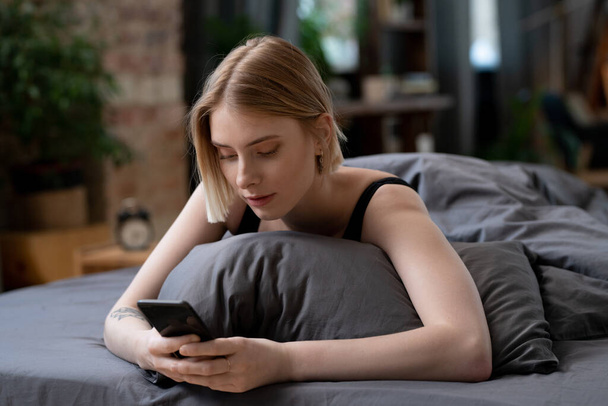 Mladá blond žena s chytrým telefonem rolování přes on-line zprávy nebo surfování v sociálních sítích v dopoledních hodinách při relaxaci na posteli - Fotografie, Obrázek