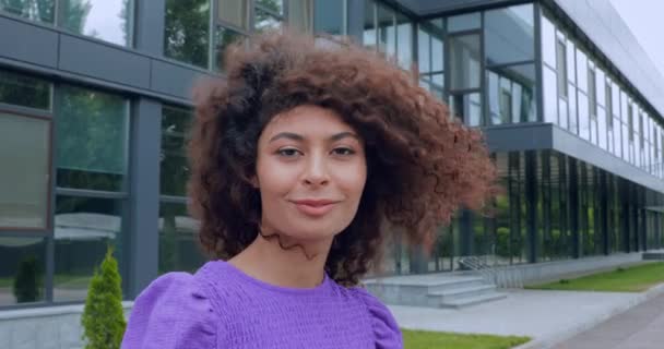 wesoła kobieta uśmiecha się i dostosowuje kręcone włosy w pobliżu budynku  - Materiał filmowy, wideo