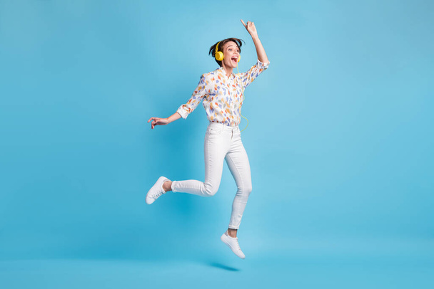 Full size photo of young girl happy excited jump listen music earphones show finger empty space isolated over blue color background - 写真・画像