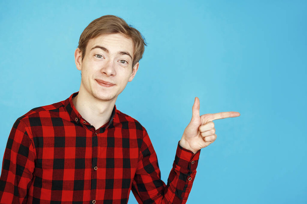 young male teenager in red shirt on blue background points finger at copy space - Foto, afbeelding