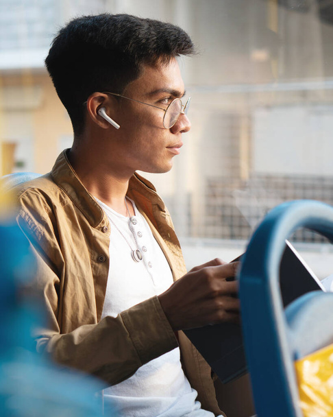 jonge Spaanse man met een bril, het lezen van een boek tijdens het gebruik van de bus naar de universiteit, het luisteren naar muziek op zijn koptelefoon - Foto, afbeelding