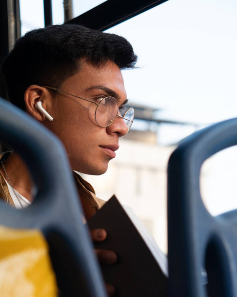 jonge Spaanse man met een bril, het lezen van een boek tijdens het gebruik van de bus om naar de universiteit te gaan, het luisteren naar muziek op zijn koptelefoon - Foto, afbeelding