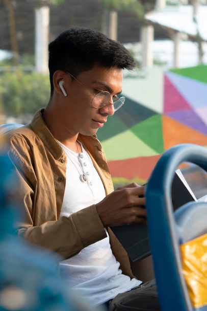 jonge Spaanse man met een bril, het lezen van een boek tijdens het gebruik van de bus om naar het werk te gaan, het luisteren naar muziek op zijn koptelefoon - Foto, afbeelding