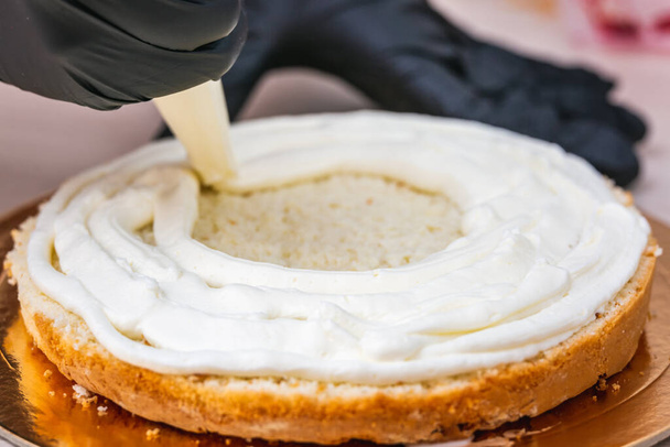 Γυναικείο χέρι σε μαύρο γάντι συμπιέζει την κρέμα σε ένα cheesecake με καρύδα. - Φωτογραφία, εικόνα