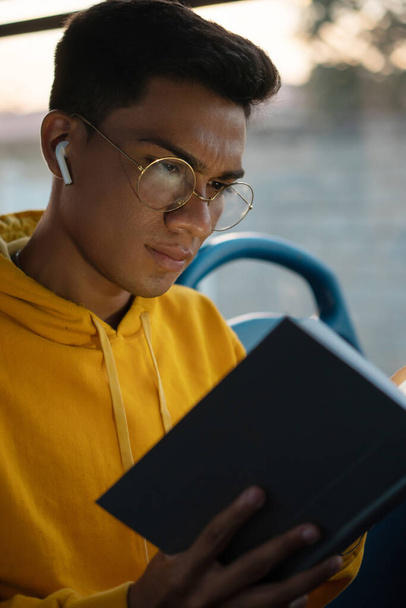 young hispanic man with glasses, reading a book while using the bus to go to university, listening to music on his headphones - Photo, Image