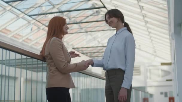 Medium shot van twee blanke zakenvrouwen die elegante kantoorkleding dragen die samen staan in de business center hall, pratend en handen schudden - Video
