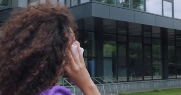 curly young woman talking on smartphone near building  - Footage, Video
