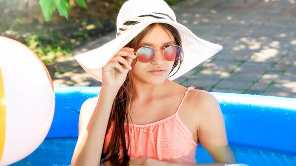 Улыбающаяся девочка-подросток в летней шляпе и стильных солнцезащитных очках расслабляется в плавательном бассейне на заднем дворе дома. Концепция счастливого летнего отдыха и отпуска - Фото, изображение