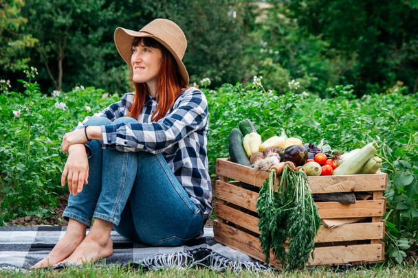 Piękna kobieta rolnik siedzi na kocu w pobliżu świeżych warzyw organicznych w drewnianym pudełku na tle ogrodu warzywnego. Kopiowanie, puste miejsce na tekst. - Zdjęcie, obraz