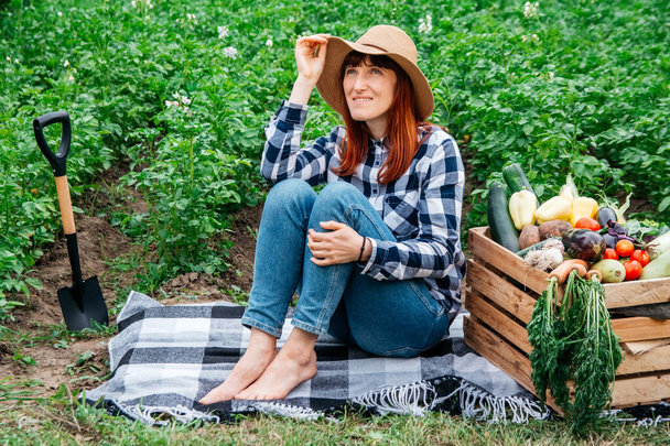 Linda agricultora com uma pá sentada em um cobertor perto de legumes orgânicos frescos em uma caixa de madeira no fundo de uma horta. Copiar, espaço vazio para texto. - Foto, Imagem