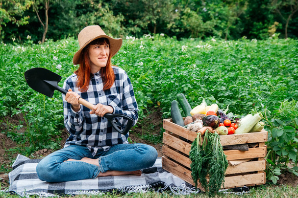 Belle agricultrice avec une pelle assise sur une couverture près de légumes biologiques frais dans une boîte en bois sur le fond d'un potager. Copier, espace vide pour le texte. - Photo, image