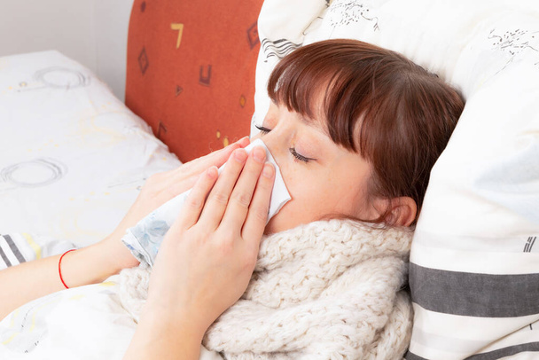 Μια νεαρή όμορφη κοπέλα είναι άρρωστη στο κρεβάτι και φυσάει τη μύτη της σε ένα μαντήλι. - Φωτογραφία, εικόνα