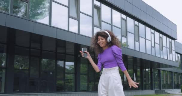 mujer feliz escuchando música en auriculares, bailando con smartphone afuera  - Metraje, vídeo