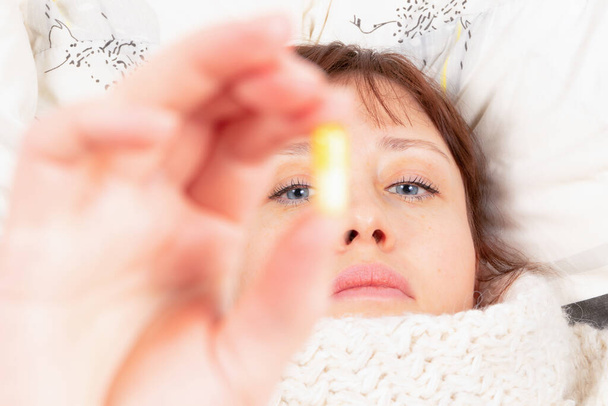 una joven bonita envuelta en una bufanda está enferma en casa, se acuesta en la cama y sostiene una cápsula con medicina en la mano - Foto, Imagen