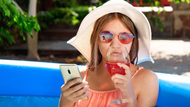 Belle fille souriante buvant un cocktail et utilisant un smartphone tout en se relaxant dans la piscine gonflable - Photo, image