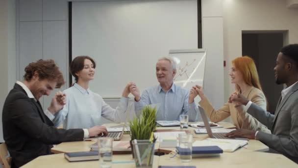 Mediana toma de cinco diversos colegas de negocios sentados juntos en el escritorio durante la reunión, tomados de la mano el uno del otro y haciendo golpe de aire - Metraje, vídeo