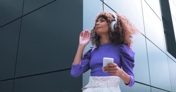 curly woman listening music in headphones and holding smartphone outside  - Footage, Video