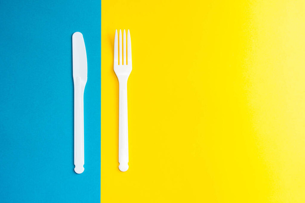 Tenedor y cuchillo plástico blanco sobre fondo amarillo y azul. Utensil de cocina. Vista superior. Estilo minimalista. Copiar, espacio vacío para texto. - Foto, Imagen