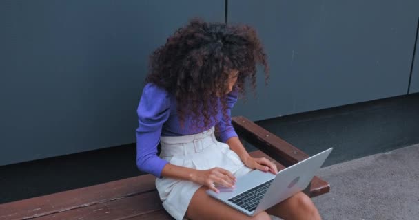 mujer rizada usando el ordenador portátil y tomando la tarjeta de crédito mientras está sentado en el banco  - Metraje, vídeo