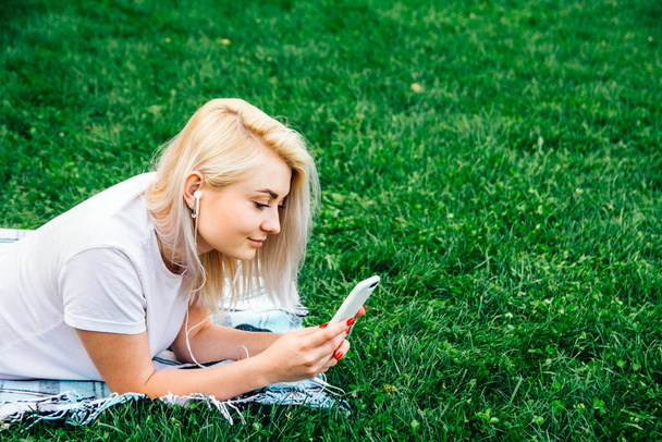 Beautiful blonde woman in headphones and smartphone in hands listens to music lying on green background. Lifestyle concept. Copy, empty space for text. - Photo, Image