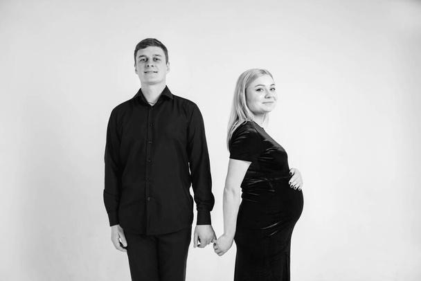 Mulher grávida e seu marido de mãos dadas em roupas pretas em um fundo branco. Foto em preto e branco. Belo casal. Copiar, espaço vazio para texto. - Foto, Imagem
