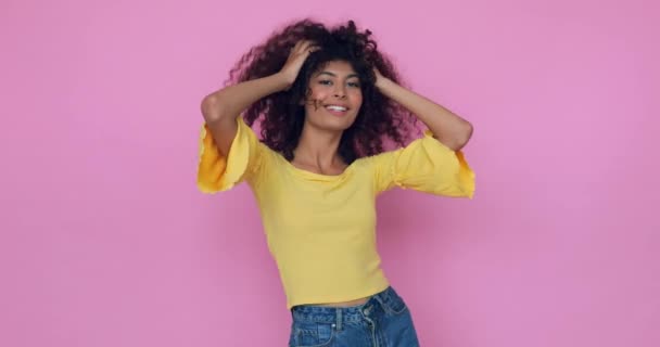 joyeux jeune modèle posant et ajustant les cheveux bouclés isolés sur rose  - Séquence, vidéo