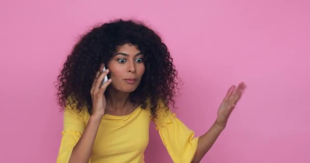 mujer joven insatisfecha haciendo gestos mientras habla en un teléfono inteligente aislado en rosa - Metraje, vídeo