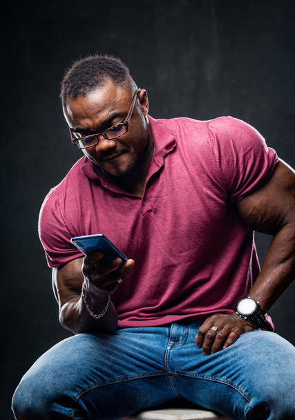 Smiling cheerful young black man poses with phone in hands over black background. African american in pink shirt. - Foto, Imagen