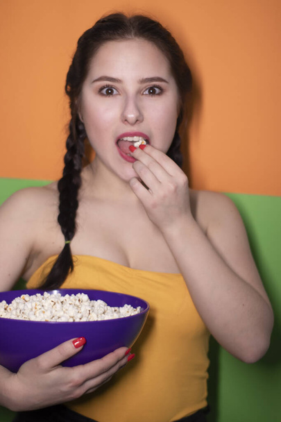 charming brunette woman in yellow top holding a purple bowl with popcorn on bright green and orange background. movie concept. cinema theme - Фото, зображення